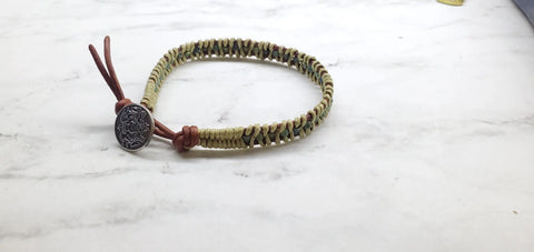 The Alexa Martha Designs Leather Seed Bead Rattlesnake Tail Weave Bracelet features elegant beige and green braids, brown leather ends, and a round Tree of Life closure, beautifully showcased on a white marble surface.