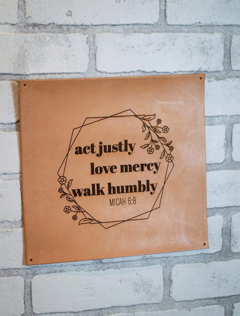 The Urban Southern Act Justly Leather Sign features floral designs and the text: act justly, love mercy, walk humbly, Micah 6:8. This handcrafted wall decor is displayed on a white brick wall.