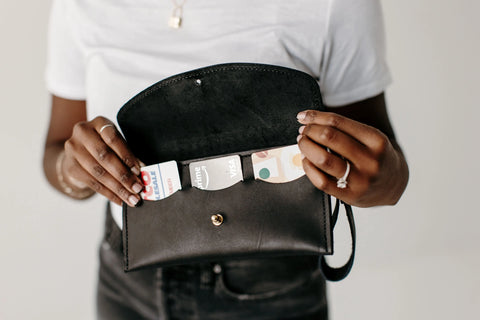 A person wearing a white shirt and rings holds open a black purse, revealing credit cards inside the Urban Southern Ruby Wristlet Wallet, handcrafted in the USA from full-grain leather.