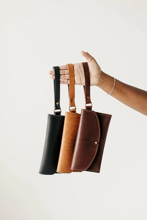 A hand displays three Urban Southern Ruby Wristlet Wallets—black, tan, and brown—each handcrafted in the USA with wrist straps and visible stitching, set against a plain light background.