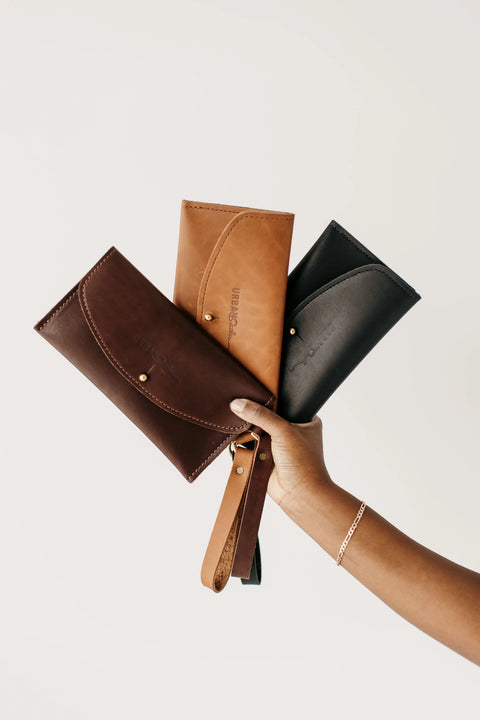 A hand displays three Urban Southern Ruby Wristlet Wallets in brown, tan, and black. Each full-grain leather wallet features a wrist strap and button closure, highlighting quality craftsmanship against a plain light background.