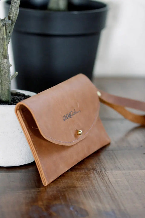 The Ruby Wristlet Wallet by Urban Southern, crafted from brown full-grain leather with a button clasp, sits on a wooden surface beside a potted plant in a white and black pot, highlighting quality US handcrafted craftsmanship.
