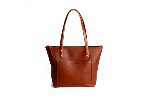 The Urban Southern Urban Zipper Tote is a brown full-grain leather tote bag with double handles, front pocket, and zipper closure, shown against a plain white background.