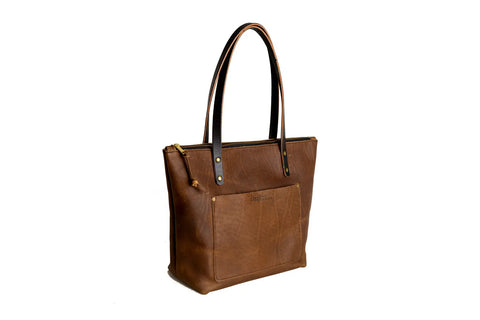 The Urban Southern Urban Zipper Tote is a brown full-grain leather bag with two long handles, a front pocket, and a zipper closure, shown on a white background.