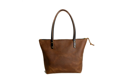 The Urban Southern Urban Zipper Tote is a brown full-grain leather tote with two dark handles and a zipper closure, shown against a white background.