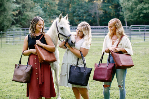 Three women stand in a grassy field, each holding an Urban Southern Urban Zipper Tote in various colors. A white horse nuzzles the center woman, with trees and a fence visible in the background.