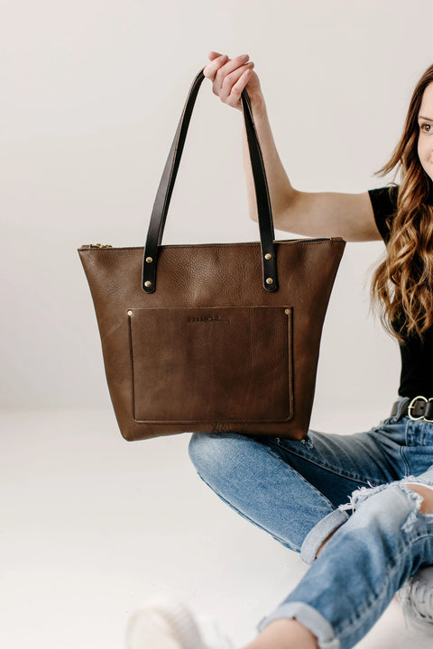 A woman in a black shirt and ripped jeans sits on the floor, holding up the Urban Southern Urban Zipper Tote with black handles against a plain white background.