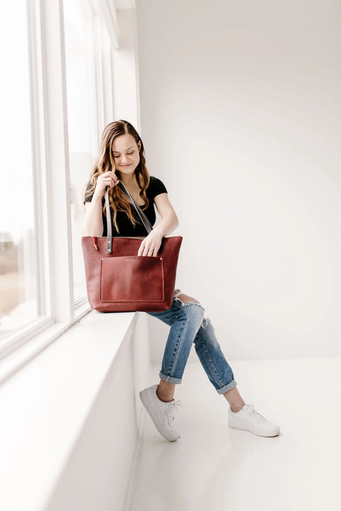 A woman in a black t-shirt and ripped jeans sits on a windowsill in a bright room, looking into the Urban Southern Urban Zipper Tote in red leather. She wears white sneakers and smiles slightly.