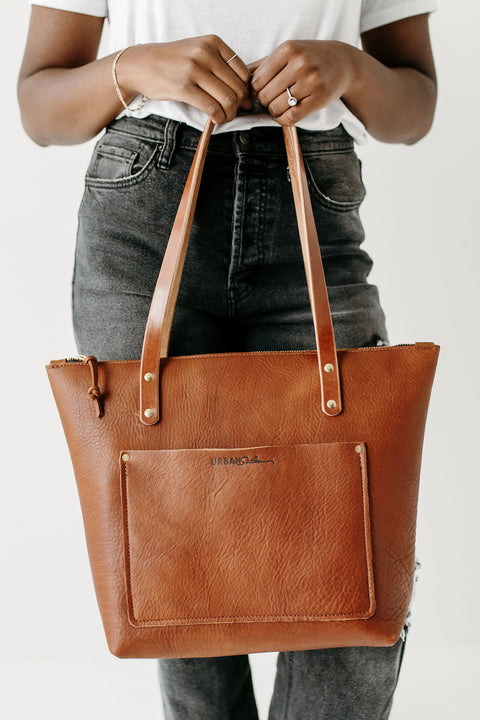 A person in a white shirt and black jeans holds the Urban Southern Urban Zipper Tote, an American-made full-grain leather bag with double handles, front pocket, gold hardware, and a subtle logo.