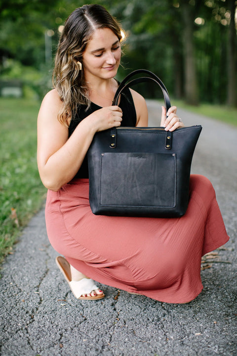 A woman with wavy hair kneels on a path, holding the Urban Southern Urban Zipper Tote. She wears a black sleeveless top and long coral skirt, surrounded by green grass and trees in the background.