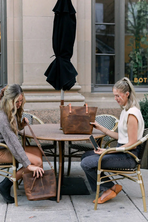 Two women sit at an outdoor café table with Urban Southern Urban Zipper Tote bags; one looks into her full-grain leather bag while the other holds a phone. A closed black umbrella rests on the table, with a building in the background.