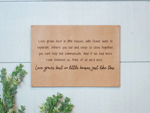 The Urban Southern Love Grows Best Leather Sign, featuring black handwritten text about love thriving in small homes, hangs on a light gray wood wall amid green leafy plants.