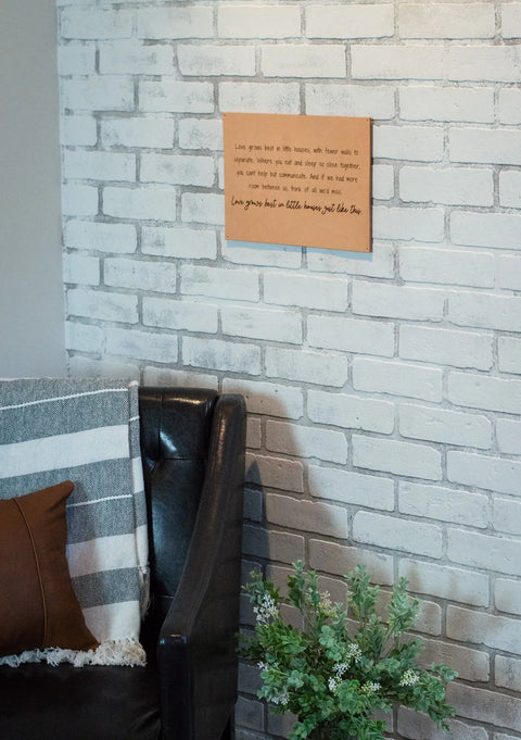 A cozy corner features a black leather chair, brown pillow, striped blanket, and green plant. Above, the Urban Southern Love Grows Best leather sign adds a rustic touch to the white brick wall.