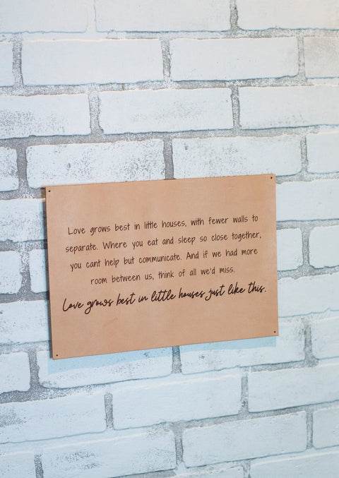 The Urban Southern Love Grows Best Leather Sign, featuring engraved cursive and printed text, brings rustic charm to any space with the phrase: “Love grows best in little houses just like this.” Mounted on a white-painted brick wall.