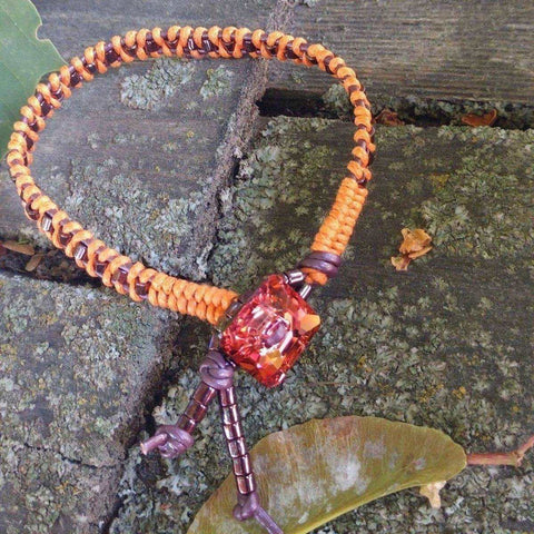 The Orange Purple Leather Beaded Rattlesnake Tail Weave Bracelet by Alexa Martha Designs features a vibrant centerpiece and is shown on rustic wood with moss and fallen leaves.