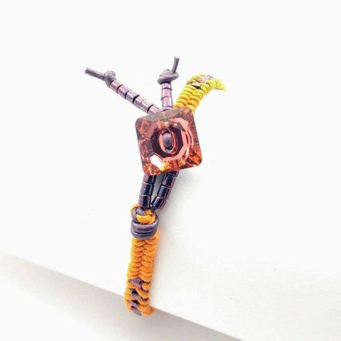 The Orange Purple Leather Beaded Rattlesnake Tail Weave Bracelet by Alexa Martha Designs features orange and yellow cord, purple beads, and a large square orange gemstone centerpiece, beautifully displayed on a white background.