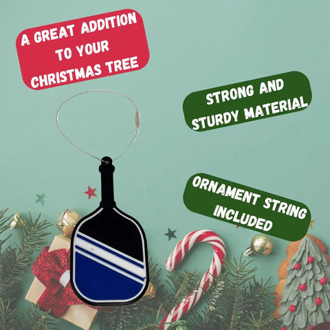 The Chatelet Manufacturing Pickleball Paddle Christmas Ornament, made in the USA, features blue and white stripes and hangs by a silver string over pine branches and gifts—highlighted for its strong design and ideal as a holiday pickleball gift.