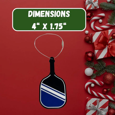 The Chatelet Manufacturing Pickleball Paddle Christmas Ornament, made in the USA, hangs on a red background with wrapped gifts and candy canes. Above, DIMENSIONS 4 X 1.75 appears in white on a green box. Perfect holiday ornament for pickleball fans!.
