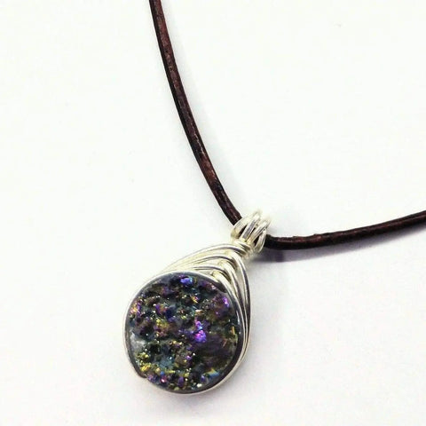 The Wire Wrapped Rainbow Druzy Necklace by Alexa Martha Designs features a round, multicolored titanium druzy agate pendant in a sterling silver herringbone wrap on a dark brown leather cord, shown against a white background.