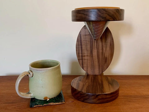 A green ceramic mug on a coaster sits beside The Rounded Pour Over by Shop Cat Woodworks—a handcrafted coffee kit with a reusable cone filter—both resting on a wooden surface against a plain light wall.