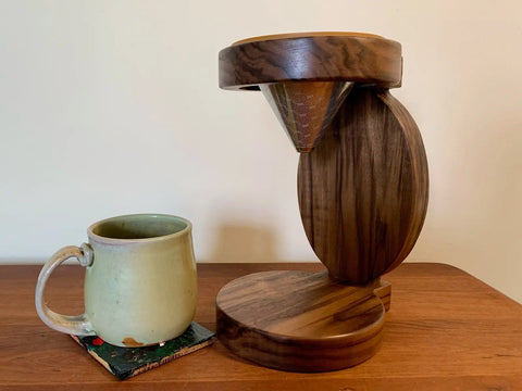 The Rounded Pour Over from Shop Cat Woodworks, featuring a reusable cone filter, sits beside a ceramic mug on a coaster on a wooden table against a light-colored wall.