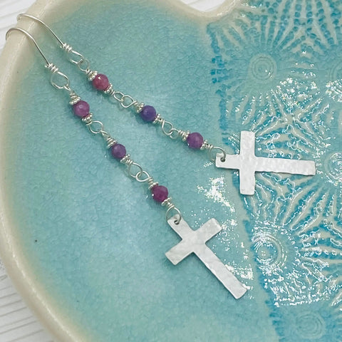 The JANECKA Ruby Rosary Earrings by Janecka, featuring hand-forged silver crosses with purple and pink beaded chains, are displayed on a blue textured ceramic dish.