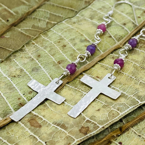 The Janecka Ruby Rosary Earrings feature hand-forged silver crosses with pink and purple beads, displayed on dried leaves—a perfect handmade gift for lovers of unique jewelry or a stylish July birthstone-inspired accessory.