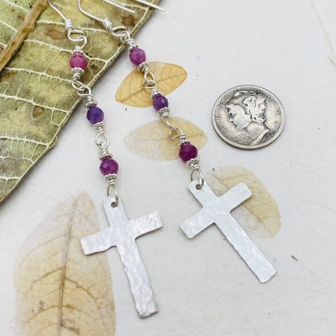 The Janecka Ruby Rosary Earrings feature hand-forged crosses and purple beads—ideal for handmade jewelry enthusiasts. Shown beside a US dime for scale, theyre displayed on dried leaves with a lacy leaf skeleton.