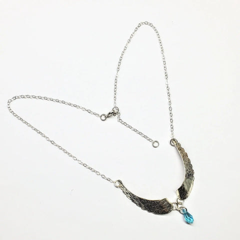 The Handmade Silver Forged Sculpted Angel Wings Necklace by Alexa Martha Designs features two curved, textured wing pendants with a small blue Swarovski crystal charm, elegantly presented on a white background.