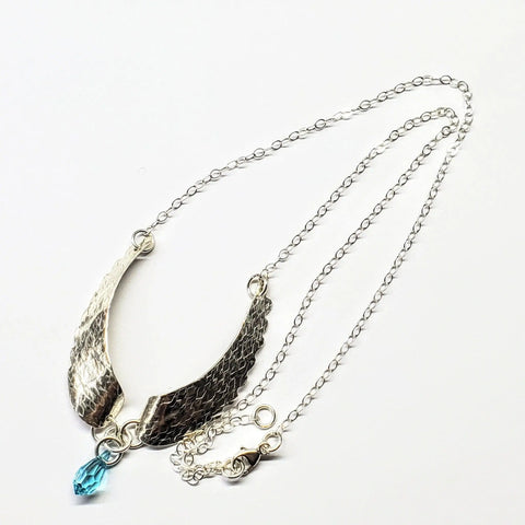 The Handmade Silver Forged Sculpted Angel Wings Necklace by Alexa Martha Designs features a textured wing-shaped pendant with a blue bead, elegantly displayed on a white background.