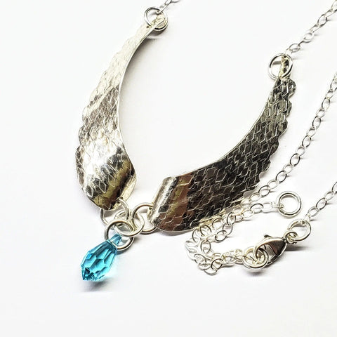 The Handmade Silver Forged Sculpted Angel Wings Necklace by Alexa Martha Designs showcases textured wing-shaped pendants and a faceted blue teardrop charm, elegantly displayed on a white background.
