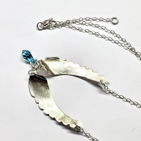 The Alexa Martha Designs Handmade Silver Forged Sculpted Angel Wings Necklace features a pendant with two open pea pods and a blue teardrop crystal, displayed on a white background with a thin chain and small clasp.