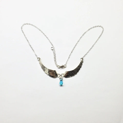 The Handmade Silver Forged Sculpted Angel Wings Necklace by Alexa Martha Designs features two wing-shaped charms and a blue teardrop pendant, elegantly displayed against a plain white background.