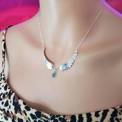 The Handmade Silver Forged Sculpted Angel Wings Necklace by Alexa Martha Designs, featuring wing-shaped pendants and a blue crystal drop, is shown on a mannequin in a leopard-print top against a pink background.