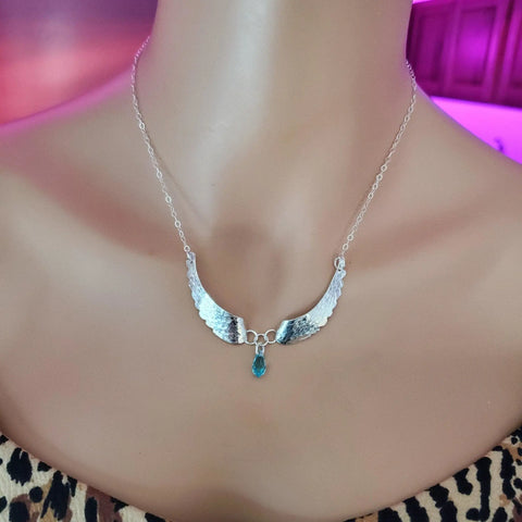 The Handmade Silver Forged Sculpted Angel Wings Necklace by Alexa Martha Designs, featuring a blue teardrop gemstone, shines on a mannequin in a leopard print top against a pink and purple background.