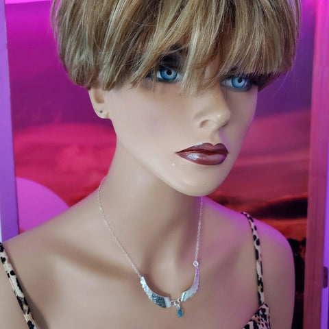 A mannequin with short blonde hair, blue eyes, and dark lipstick models the Alexa Martha Designs Handmade Silver Forged Sculpted Angel Wings Necklace with a blue pendant and a leopard print top against a pink and purple background.