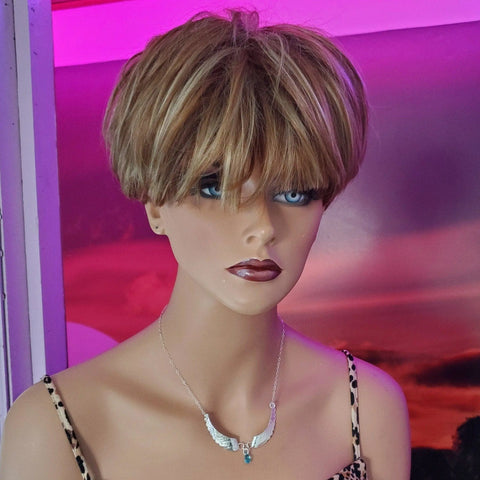 A light-skinned mannequin with short blonde and brown hair and blue eyes wears the Handmade Silver Forged Sculpted Angel Wings Necklace by Alexa Martha Designs over a leopard-print top, against a vibrant pink and purple sunset background.