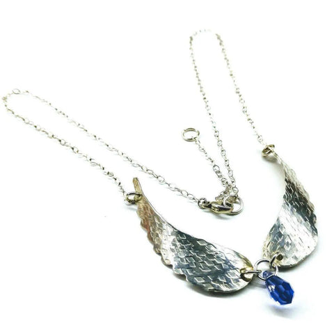 The Handmade Silver Forged Sculpted Angel Wings Necklace by Alexa Martha Designs features two textured sterling silver wing pendants joined by a blue Swarovski crystal on a delicate chain.