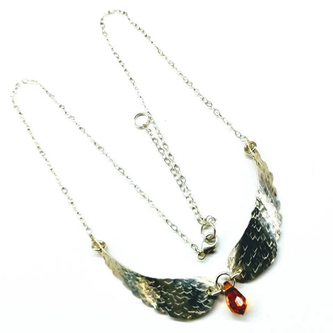 The Handmade Silver Forged Sculpted Angel Wings Necklace by Alexa Martha Designs features wing-shaped metal pendants with a textured pattern and a central orange-red teardrop gemstone.