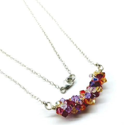 The Silver Multi Color Spring Blossom Crystal Necklace by Alexa Martha Designs features a sterling silver chain with a curved cluster of faceted red, purple, orange, and yellow beads—a vibrant handmade piece displayed on white.
