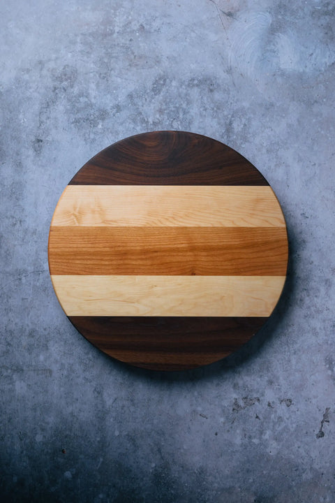 The Lazy Susan by Montana Block is a handcrafted round wooden board featuring alternating light and dark wood grain stripes, displayed on a textured gray concrete surface.