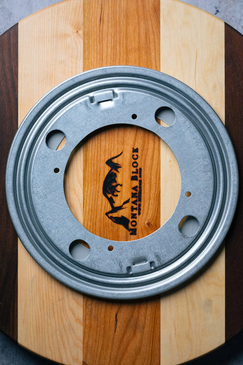 The Lazy Susan by Montana Block features a round metal plate with holes on a handcrafted solid wood striped cutting board, showcasing the brands dinosaur logo and Montana Block wording.