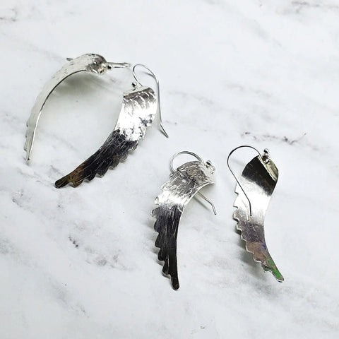 Sterling Silver Hammer Pattern Sculpted Angel Wing Earrings by Alexa Martha Designs, featuring a textured, curved design, are displayed on a white marble surface.