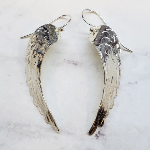 The Sterling Silver Hammer Pattern Sculpted Angel Wing Earrings by Alexa Martha Designs are handcrafted and displayed on white marble, featuring a mirrored feather-like design with detailed hammered texture.
