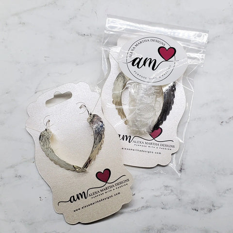Two pairs of Sterling Silver Hammer Pattern Sculpted Angel Wing Earrings by Alexa Martha Designs are shown on decorative cards, one in a clear plastic bag and one displayed on a white marble surface. Elegant, handmade jewelry.