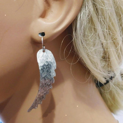 A close-up shows a person wearing Alexa Martha Designs Sterling Silver Hammer Pattern Sculpted Angel Wing Earrings. The textured, handmade design stands out against blonde hair in the background.