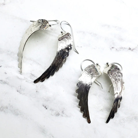 Sterling Silver Hammer Pattern Sculpted Angel Wing Earrings by Alexa Martha Designs, featuring textured details, are displayed on a white marble surface.