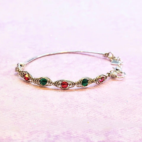 The Limited Edition Silver Wire Wrapped Red Green Christmas Holiday Tube Bracelet by Alexa Martha Designs features alternating red and green beads in sterling silver, beautifully displayed on a light pink textured surface—ideal for festive wear.