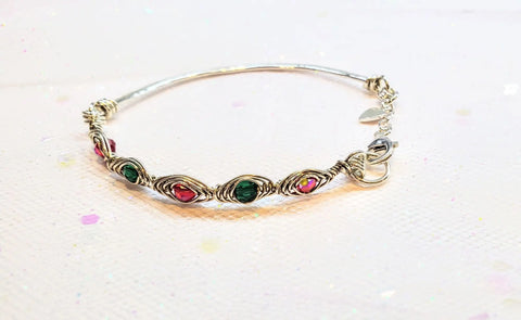 The Limited Edition Silver Wire Wrapped Red Green Christmas Holiday Tube Bracelet by Alexa Martha Designs is a handmade sterling silver piece with alternating red and green gemstones, a heart-shaped clasp, and a charm on a textured background.