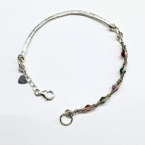 The Limited Edition Silver Wire Wrapped Red Green Christmas Holiday Tube Bracelet by Alexa Martha Designs features a textured sterling silver band, wire wrapping, colorful beads, heart charm, and lobster clasp—perfect festive bangle on white background.